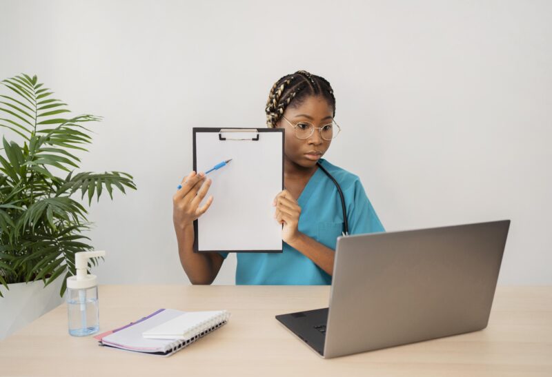 Free online health training concept—healthcare student attending virtual class with laptop, holding a clipboard, and taking notes at home